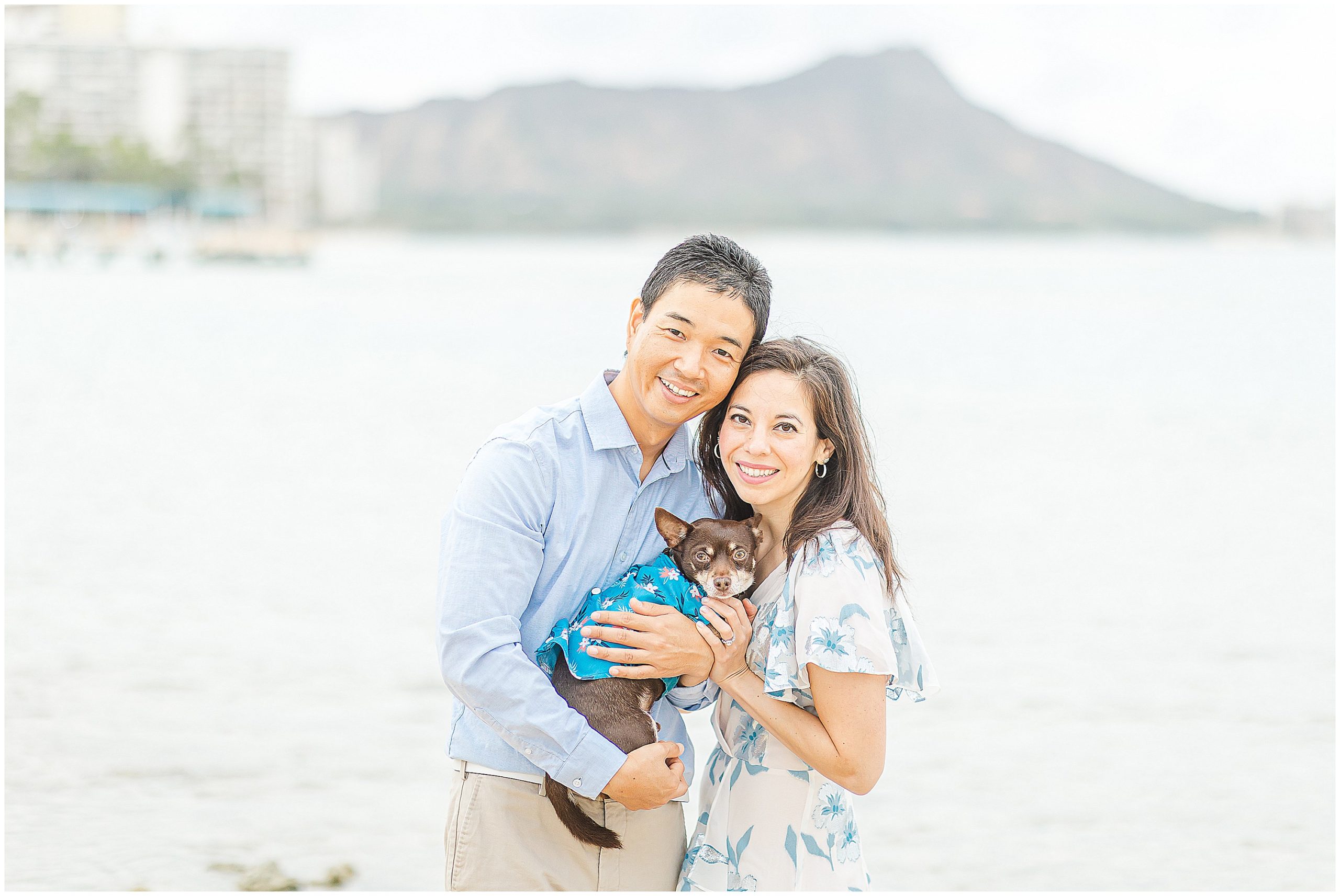 Engagement photo of a couple holding their small dog on a beach with Diamond head in the background