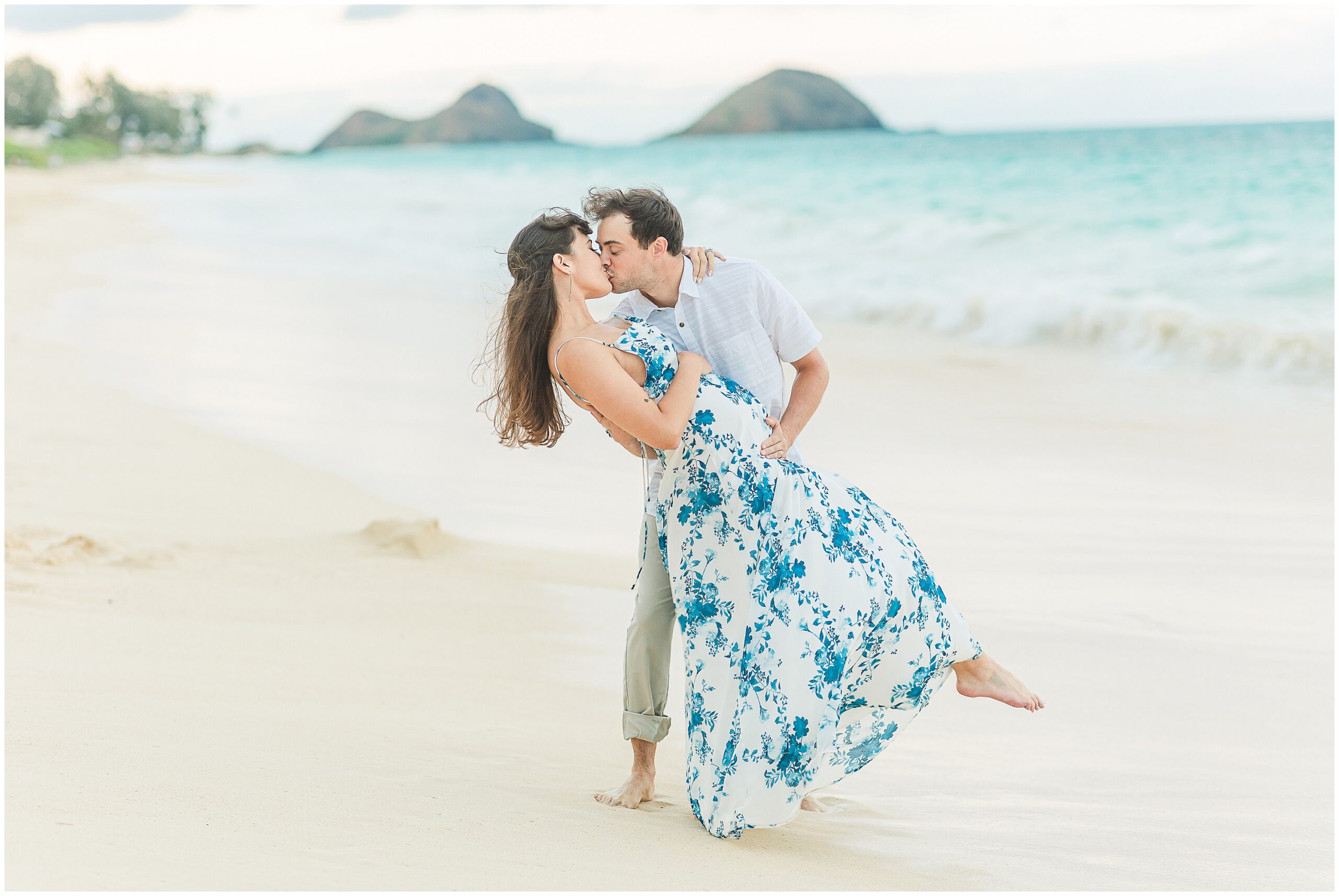 Maternity portrait of a woman in a white dress with blue florals and her husband holding her on the beach.