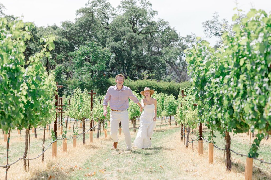 summer is the best time for photos in napa valley. this photo features a couple literally frolicking amongst the lush green vineyards in the midday sun. she's wearing a white dress and an iconic napa brim hat, while he's in a white and maroon checkered shirt. 