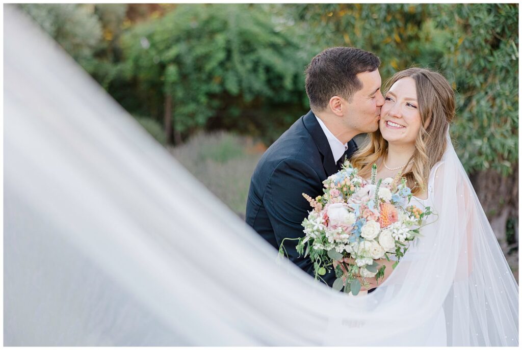 choosing the right photographer allows for this newlywed couple to have a creative portrait where the bride's veil leads to the groom kissing her on the cheek.